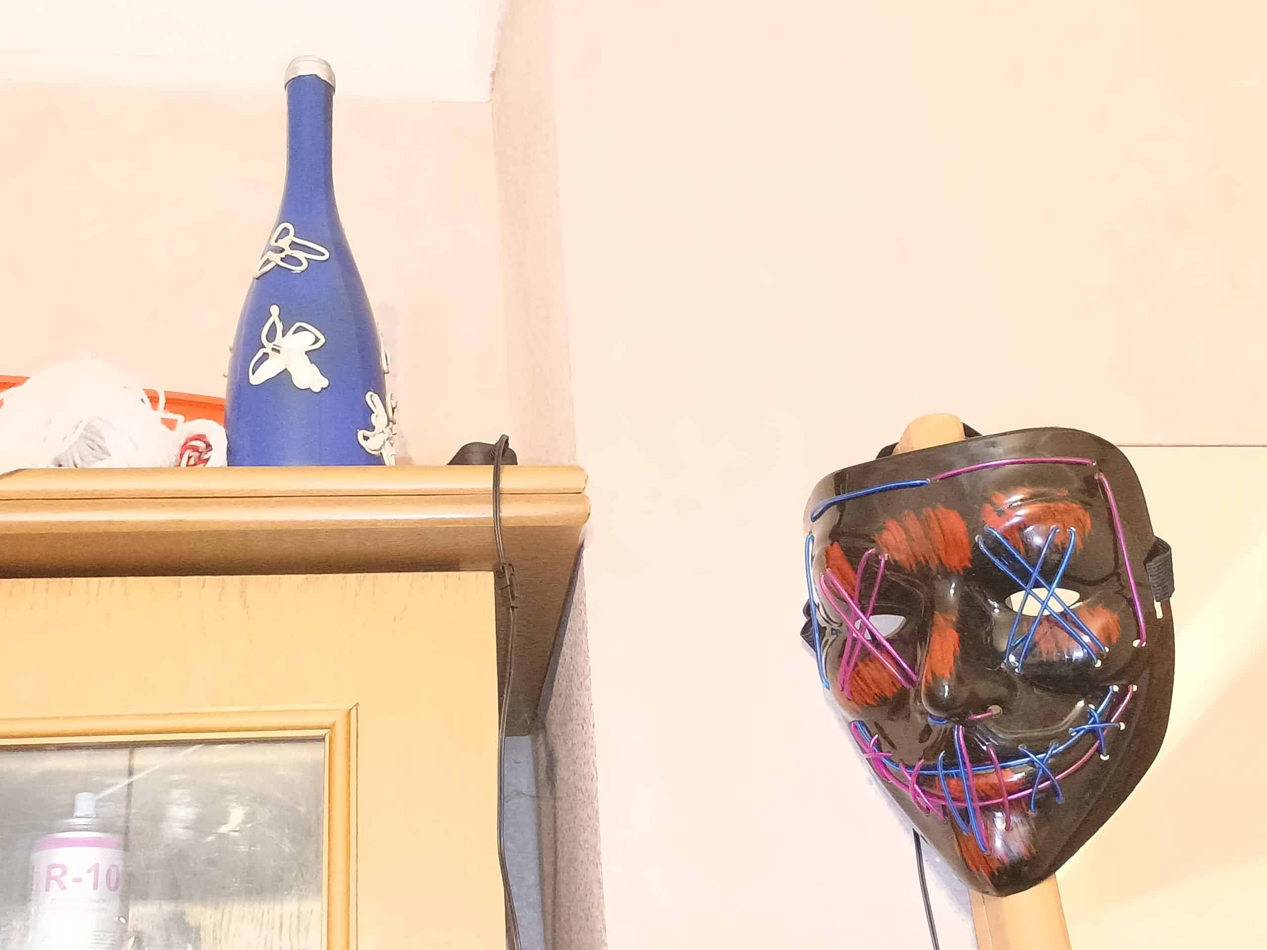 A black masquerade mask with neon stitches across the eyes and mouth, propped on a cabinet beside a blue decorative bottle. The mask symbolizes concealment and fractured identity, charm stitched together as performance, while the bottle contrasts with its still, ornamental stability.