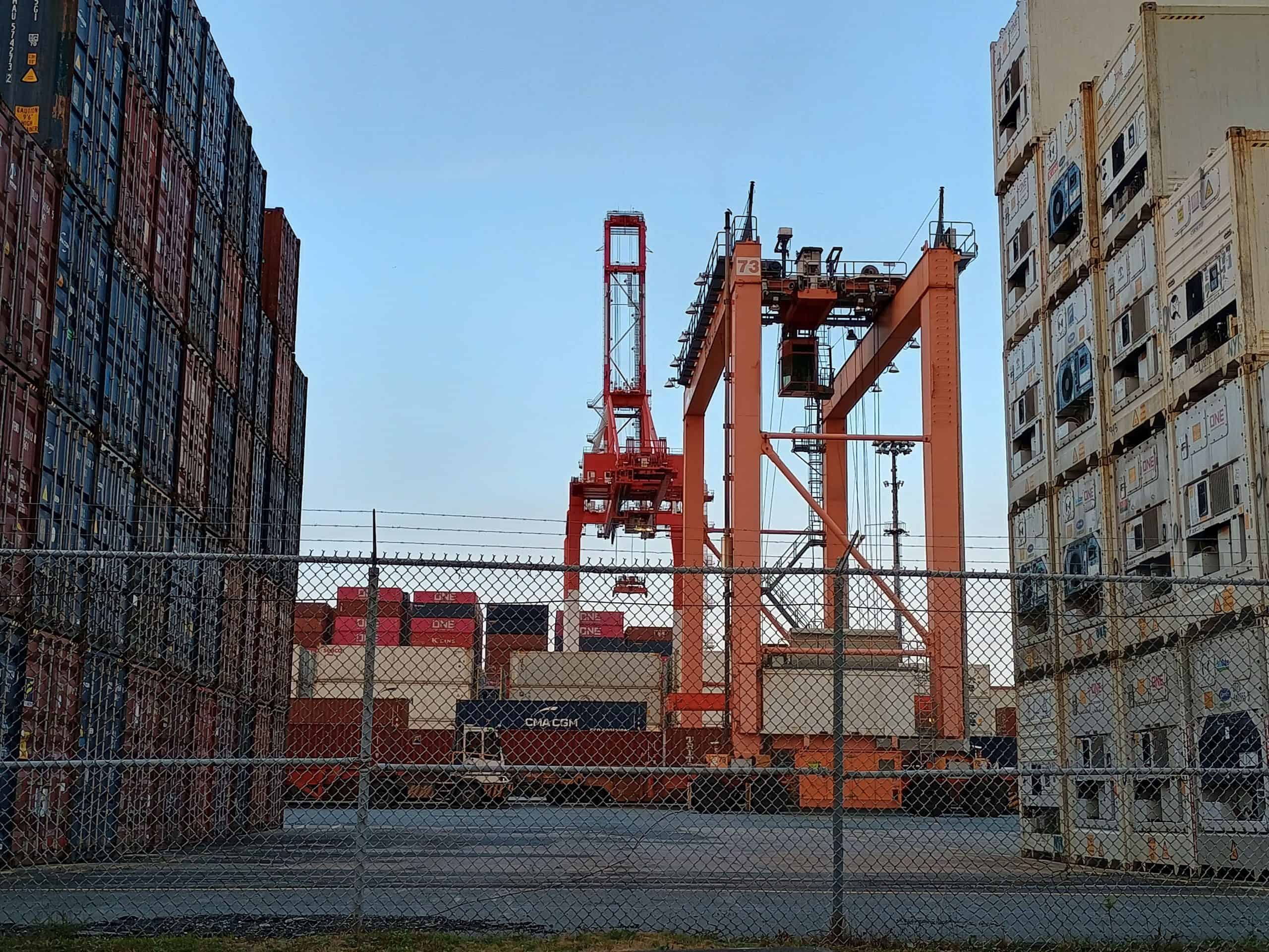 Industrial corridor framed by shipping containers, opening to cranes and open sky — representing a passage from confinement to freedom.